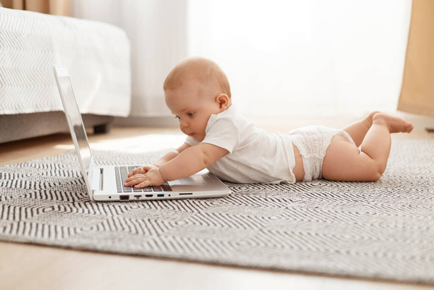 Little curious child studying modern technology while lying on floor on tummy against window, toddler using laptop at home, infant wearing white t shirt. - Zdjęcie, obraz