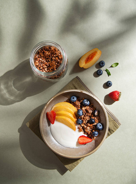 Delicious natural yogurt with homemade granola, peach, strawberry and blueberry in a bowl and jar with muesli on a green background morning shadows. Healthy and nutritious breakfast. Top view. - Photo, Image