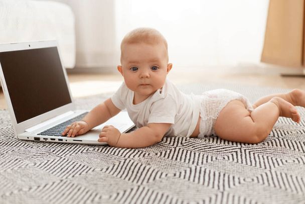 Indoor side view portrait of cute child lying on floor near laptop with blank screen and looking at camera, wearing white t shirt, posing in living room against window. - Photo, image