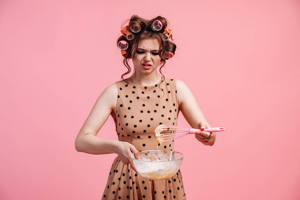 Upset woman with hair curlers on the background of an empty wall holding a plate with a whisk. Place for text, copy space. - Photo, Image