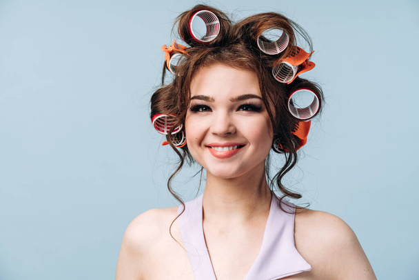 Portrait of a cute, young woman with curlers in her hair isolated on a blue background. Smiling girl in a dress, close-up, copy space, place for text. - Fotó, kép
