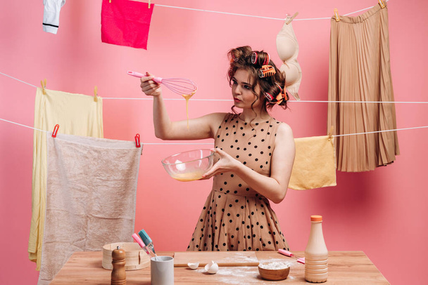 Uma mulher a fazer tarefas domésticas. Contra o pano de fundo de roupas penduradas, uma mulher com encrespadores prepara comida. - Foto, Imagem