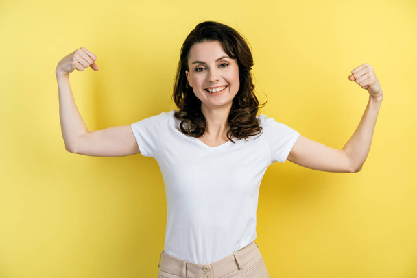 The pretty female model keeps fit and healthy, raises her arms and shows off her muscles, is proud of her achievements in the gym, and smiles broadly. Cute girl posing on a yellow wall background. - Fotografie, Obrázek