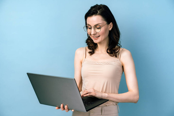 Lächelnde Geschäftsfrau mit Brille am Laptop auf blauem Hintergrund. Mädchen läuft isoliert auf leerem Wandhintergrund, Kopierraum. - Foto, Bild