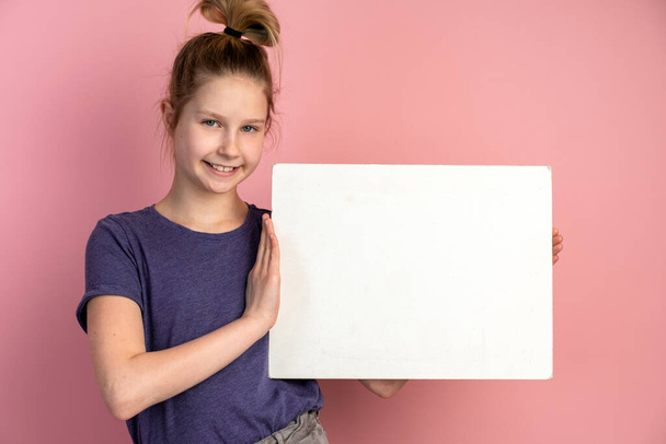 Piękna blondynka nastolatka z niebieskimi oczami trzyma białą kartkę papieru na różowym tle i uśmiecha się. Miejsce na tekst, baner, powierzchnię na napisy i reklamy. - Zdjęcie, obraz