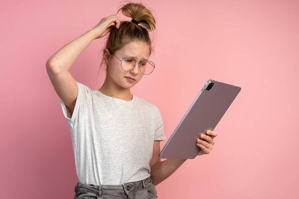 Teenage girl is studying, she is wearing glasses and a tablet in her hands. Cute little girl isolated on pink background. - Photo, Image