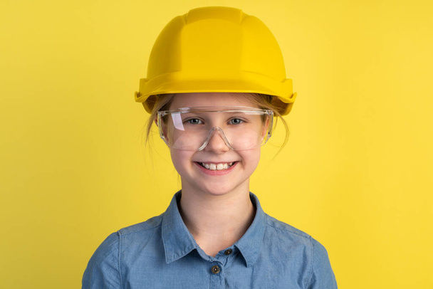 Весёлый, улыбающийся ребенок в шлеме и в строительных очках на жёлтом фоне. Девушка изолирована на пустом фоне стены. - Фото, изображение