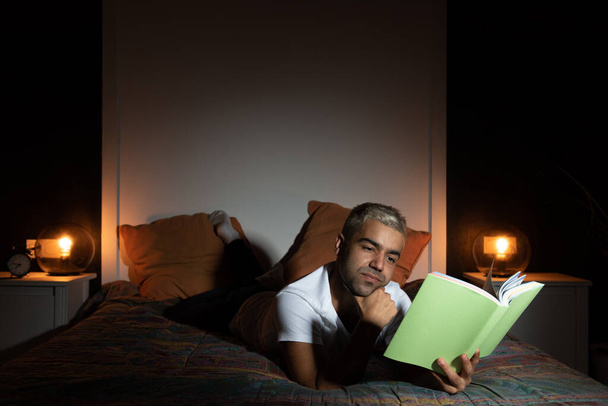 Hombre hispano leyendo un libro en el dormitorio tarde en la noche. - Foto, Imagen