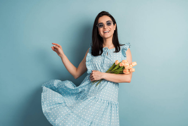 Güzel, pozitif, puantiyeli bir elbise giymiş genç bir kadın ve elinde mavi bir arka planda poz veren çiçekler. Bahar havası konsepti. - Fotoğraf, Görsel