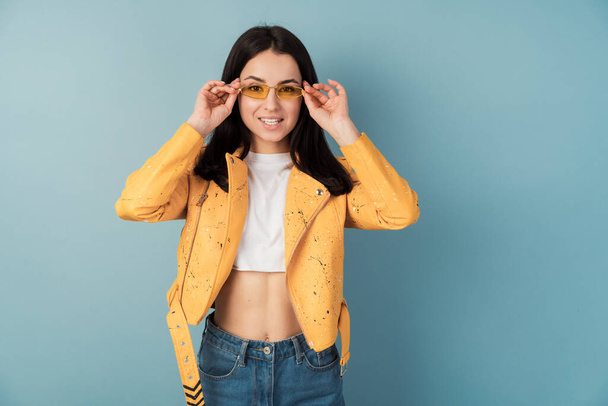 Positive girl adjusts sunglasses, she wears an orange casual jacket and blue jeans. Pretty lady on blank wall background, copy space, place for text. - Photo, Image