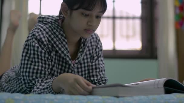 Thailändische Sekundarschülerin liest und blättert in einem Buch, während sie zu Hause auf dem Bett liegt. Bildung mit häuslicher Isolation. Freizeit und Hobbys. Teenager-Lebensstil. - Filmmaterial, Video