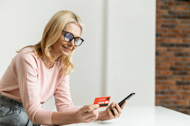A smiling middle-aged woman holds a credit card and a smartphone indoor - Photo, image