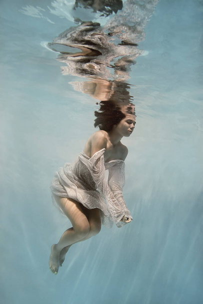 A girl in a blue dress swims underwater on a light background as if in weightlessness - Photo, image