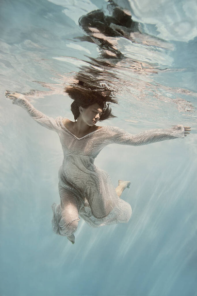 A girl in a blue dress swims underwater on a light background as if in weightlessness - 写真・画像