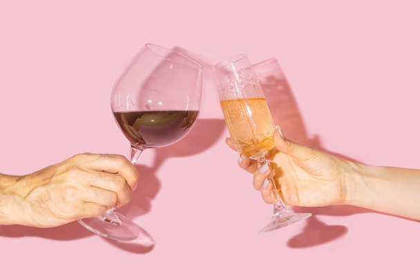 Party cheers concept. Two hands, man one holding glass of vine and girl hand holding vintage crystal glasses with lovely drinks in it against baby pink background - Photo, Image