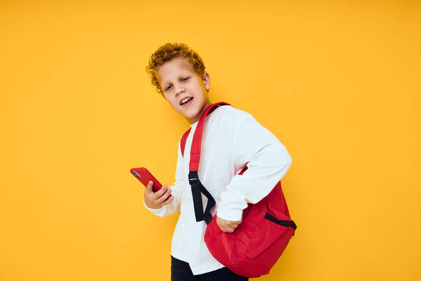 χαρούμενο σχολιαρόπαιδο σε λευκό φούτερ κινητό τηλέφωνο κόκκινο σακίδιο κίτρινο φόντο - Φωτογραφία, εικόνα