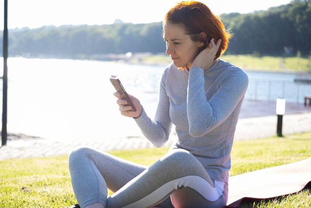 slim ginger woman in sport clothes using mobile phone at park. Health, wellness, fitness lifestyle. - Photo, Image