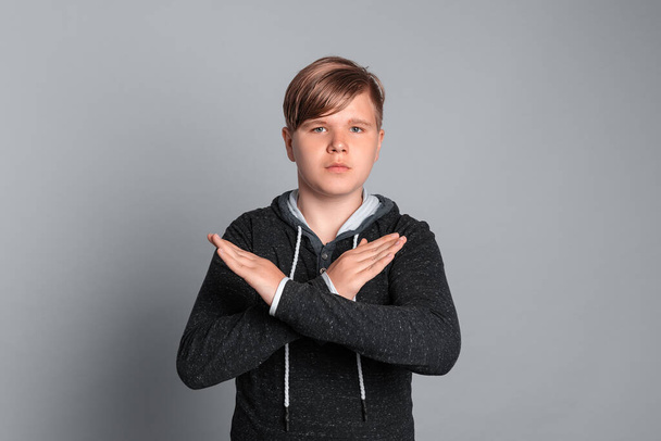 Portret poważnego nastolatka robiącego znak X ramionami, by przestać robić coś z negatywnym i poważnym wyrazem twarzy, stojącego nad szarym tłem. Stój, nie, odsuń się - Zdjęcie, obraz