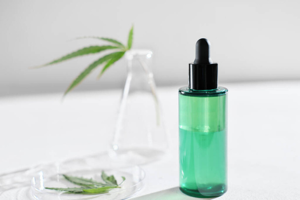 cannabis face serum in green bottle and hemp leaves in laboratory . petri dish and glassware on lab table. dermatology concept, alternative treatment. front view. - Photo, Image