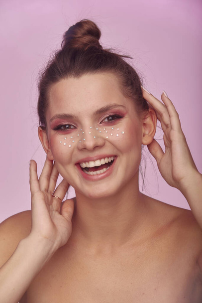 Young woman face expression with fashion creative make up. Clean fresh skin and bright make up with rhinestones freckles on her face posing on a bright pink background - Fotografie, Obrázek