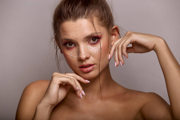 Beautiful portrait of young woman with fashion creative make up. Clean fresh skin and bright make up with rhinestones freckles on her face posing on a gray background - Photo, Image