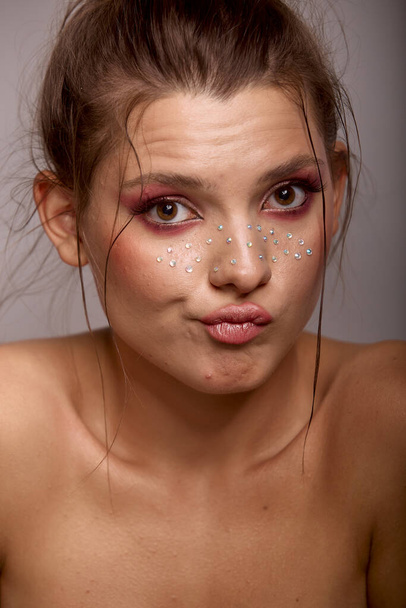 Young woman face expression with fashion creative make up. Clean fresh skin and bright make up with rhinestones freckles on her face posing on a gray background - Photo, Image