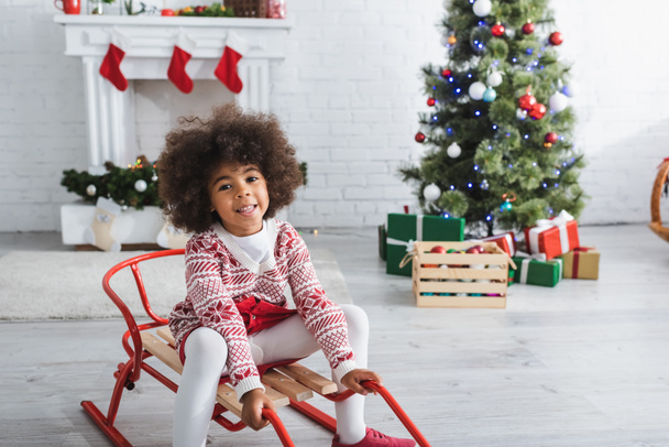 alegre niño afroamericano sentado en trineo en la sala de estar con chimenea decorada y árbol de Navidad sobre fondo borroso - Foto, Imagen