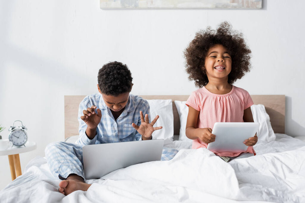 african american boy using laptop in bed near cheerful sister with digital tablet - Photo, Image