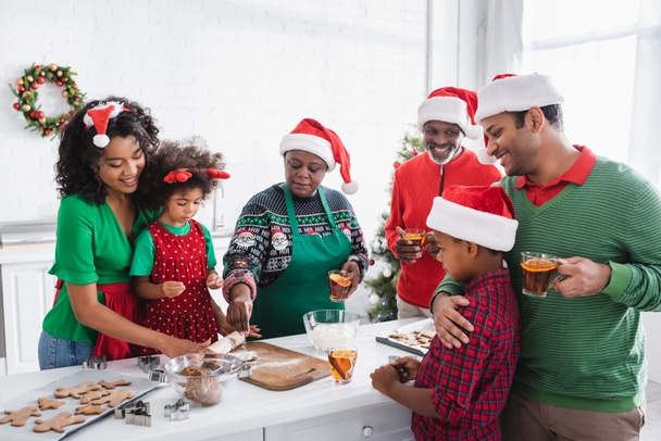 χαρούμενη αφρικάνικη αμερικανική οικογένεια πίνοντας τσάι κανέλας πορτοκάλι ενώ ετοιμάζετε χριστουγεννιάτικα μπισκότα στην κουζίνα - Φωτογραφία, εικόνα