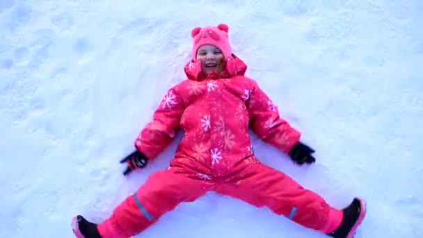 Ein junges Mädchen in gelber Jacke liegt im Schnee und bastelt einen Schneengel. - Filmmaterial, Video