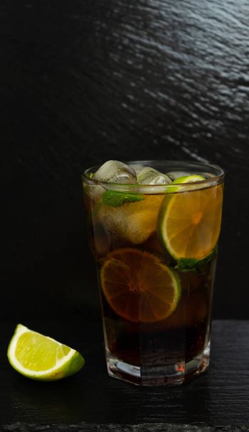 Cuba Libre cocktail with brown rum, lemon juice, cola and ice cubes, garnished with a slice of fresh lime on a modern black stone tray. Bar concept - Zdjęcie, obraz