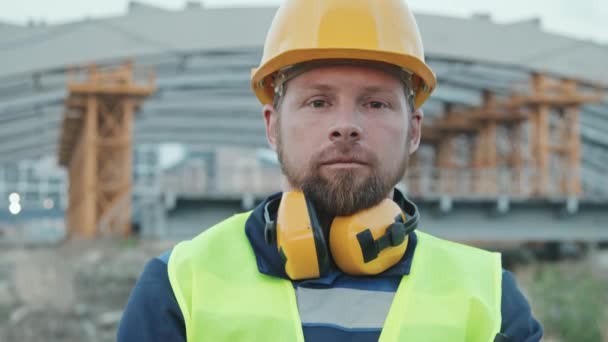 Sledování zblízka portrét vousatého stavebního pracovníka v bezpečnostní vestě a tvrdém klobouku pózujícího pro kameru na pracovišti - Záběry, video