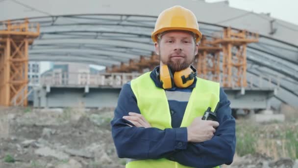 Portrait de suivi du travailleur de la construction masculin joyeux en gilet de sécurité et casque de sécurité debout avec les bras croisés et regardant la caméra à l'extérieur - Séquence, vidéo