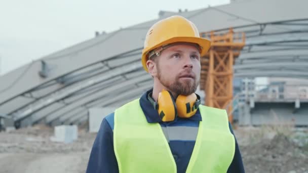 Lento seguimiento de tiro de barbudo capataz del sitio de construcción masculino en sombrero duro y chaleco de seguridad mirando a su alrededor y caminando al aire libre en el lugar de trabajo - Metraje, vídeo