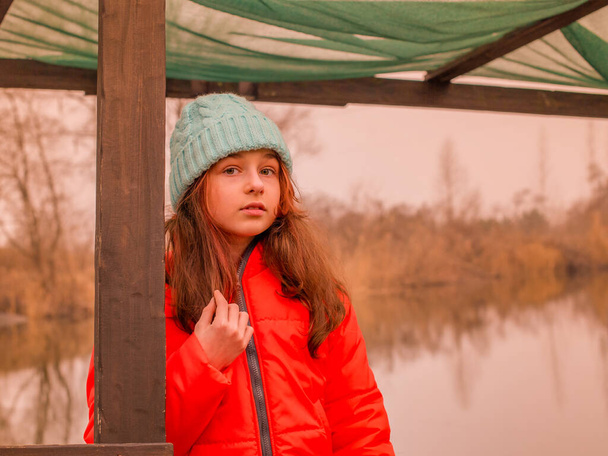 Portrait of a little girl in an orange jacket on the background of the river in the cold season. - Photo, Image