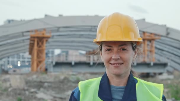 Śledzenie portret z powolnym tempie szczęśliwy pracownik budowy kobiet w kamizelce bezpieczeństwa i twardy kapelusz stoi na zewnątrz i uśmiecha się do kamery. Nieukończony budynek lub most z rusztowaniami w tle - Materiał filmowy, wideo