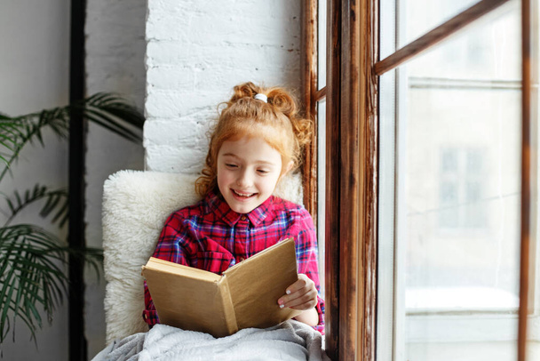 Little girl child reads a book. Child on the windowsill. Big window. Warm blanket. Distance learning. - Photo, Image