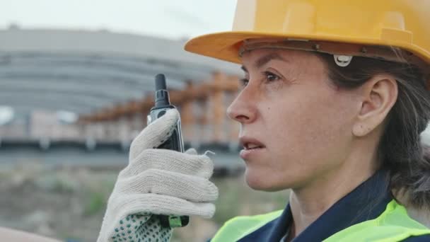 Handheld close-up schot van vrouwelijke bouwvakker in veiligheidsvest, harde hoed en handschoenen praten in walkie-talkie buiten - Video