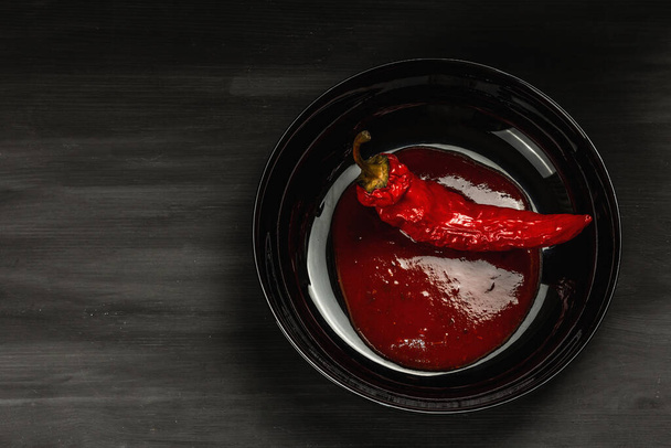 Ripe hot chili pepper sauce and whole vegetables on black wooden background. Traditional ingredient for different food, culinary backdrop, top view - Photo, Image