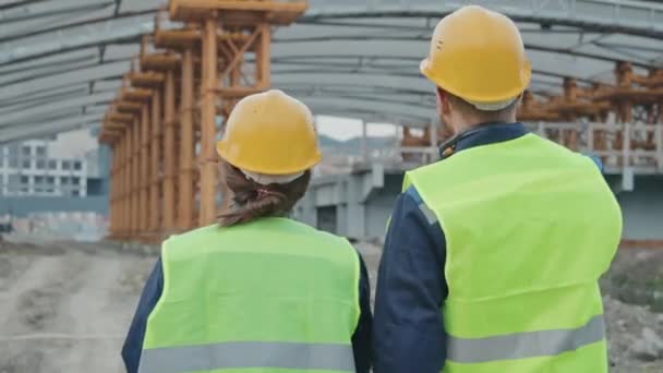 Sledování zadní pohled na mužské a ženské stavební dohlížitele v bezpečnostních vestách a tvrdých čepicích kráčející směrem k nedokončeným budovám nebo mostu s lešení a konverzace - Záběry, video
