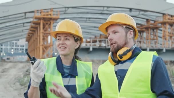 Kézi felvétel vidám női felügyelő walkie-talkie és férfi dolgozó visel biztonsági mellény és kemény kalap beszél odakint, mielőtt befejezetlen épület vagy híd állványzat - Felvétel, videó