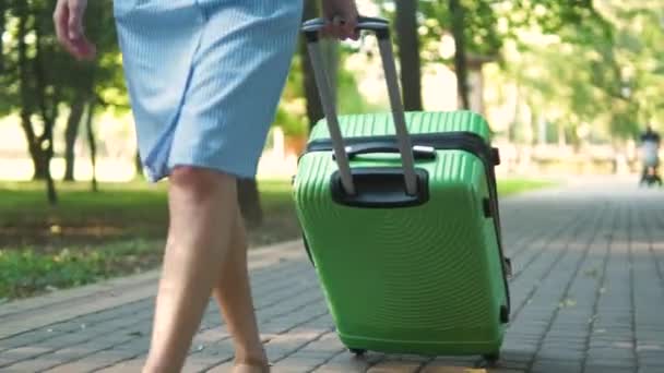 Closeup of young woman walking on city street sidewalk with green suitcase on summer day. Travelling and vacation concept. - Footage, Video