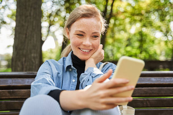 Woman sitting at the bench and using her smartphone while walking at the park - Foto, imagen