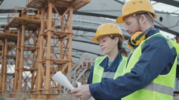 Low angle medium shot of female and male construction site workers in safety vest and hard hats standing before unfinished bridge or building with scaffolding and discussing blueprint - Footage, Video