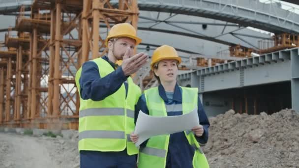 Tracking shot de ingenieros masculinos y femeninos en chaleco de seguridad y sombreros duros discutiendo planos y caminando en el sitio de construcción con andamios - Metraje, vídeo