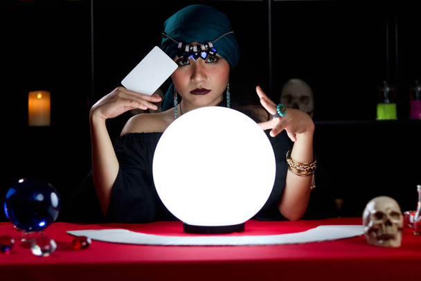 Misteriosa magnífica hermosa adivina en vestido negro sosteniendo una tarjeta y leyendo el futuro en la bola de cristal luminoso, bruja oscura lanzando un hechizo con bola mágica - Foto, Imagen