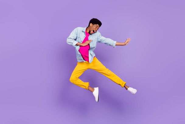 Profile photo of astonished active afro guy jump hit leg wear blue shirt pants sneakers isolated purple color background - Fotó, kép
