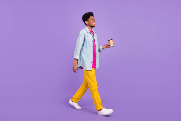 Photo of sweet funky dark skin man wear denim shirt smiling walking drinking tea looking empty space isolated purple color background - Foto, Bild