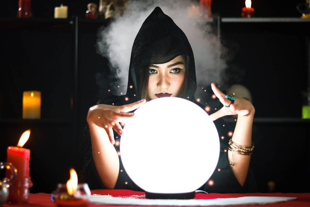Dark witch in black hood casting spell on luminous magical ball with smoke light background, mysterious magnificent beautiful woman fortune teller read future on crystal ball, scary Halloween concept - Photo, Image