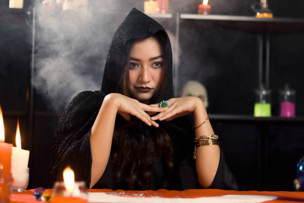 Ciemna czarownica w czarnym kapturze ustawienie przy magicznym stole z tle światła dymu, tajemniczy wspaniały piękna kobieta rzuca klątwę, przerażający pomysł Halloween - Zdjęcie, obraz